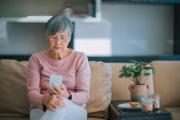 市内のアパートのリビングルームでテキストメッセージを読むスマートフォンを使用しているアジア系中国人のシニア女性 - senior adult women adult tan ストックフォトと画像