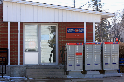Acme, Alberta, Canada- March 22, 2022:  Small town and small building that houses the Canada Post post office in Acme Alberta. Exterior of post office with three postal boxes at front entrance. Old wood house in background.