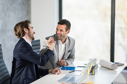 Two cheerful businessmen giving each other high-five while celebrating at work in the office.