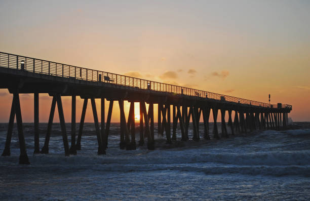 Hermosa Beach Pier at sunset.   Sun showing through pier pylons.    Rough see, clouds in the low sky.