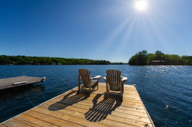 Sunrise on two empty Adirondack chairs Cottage life - Sunrise on two empty Adirondack chairs sitting on a dock on a lake in Muskoka, Ontario Canada. The sun light create long shadows on the wooden pier. rising sun chair stock pictures, royalty-free photos & images