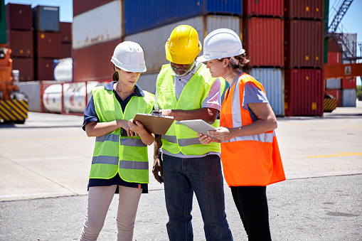 Front view of transportation manager and dock workers discussing project over clipboard and digital tablet while standing on freight terminal