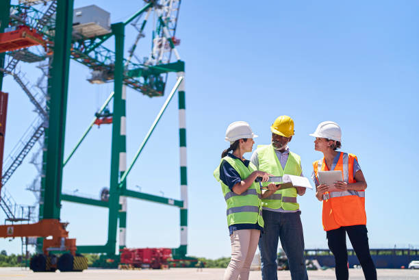 Foreman engineer and dock workers standing against harbor cargo crane while discussing work stock photo