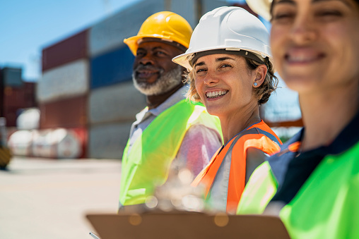 Side view of female dock worker wearing hardhat and reflective vest standing between coworkers at shipping yard while looking and smiling at the camera