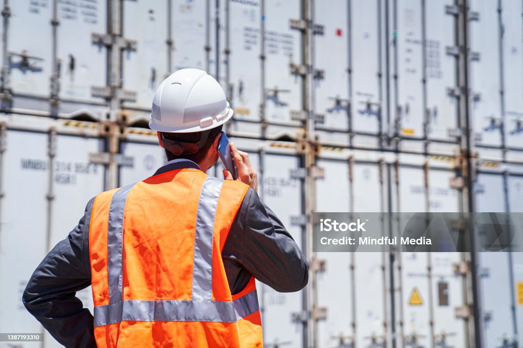 Asian american male customs broker talking on smart phone while looking at containers Rear view of asian american male customs broker wearing hardhat talking on smart phone while looking at stacks of containers during daytime Customs Official Stock Photo