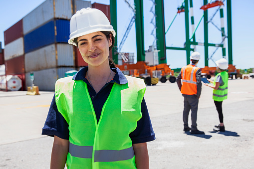 Portrait of female dock control worker looking and smiling at the camera while coworkers standing on background at shipping containers yard during daytime