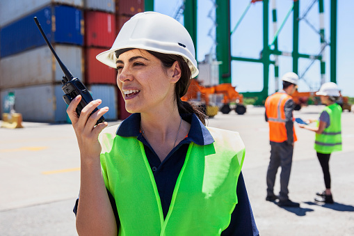 Portrait of female dock control worker talking on walkie talkie while coworkers standing on background at shipping containers yard during daytime