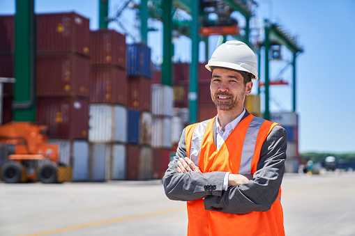Portrait of asian american male engineer wearing reflective vest and hardhat standing with arms crossed against stacks of containers at shipping container yard during daytime