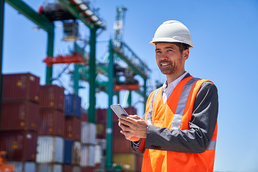 Waist up of asian american male engineer wearing hardhat and reflective vest using smart phone while standing at shipping container yard during daytime