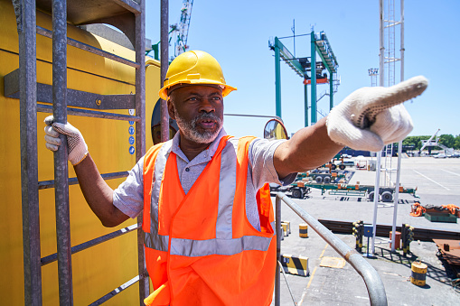 Medium shot of african american male cargo handler standing on harbor cargo crane ladder while pointing during day at shipping yard