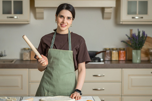 Portrait of a young brunette smiling woman in a kitchen apron with a rolling pin in her hands in a kitchen interior.