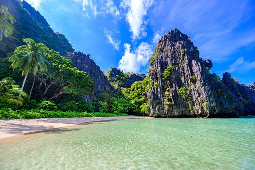 A tropical beach in Phi Phi Don Island, Thailand, with clear blue water and white sand. The beach is surrounded by green trees and rocks on one side and a mountain range on the other side. The water is a beautiful shade of turquoise and is very clear, reflecting the sky and the mountains. The sky is blue with a few clouds, creating a contrast with the green landscape. The sand is white and there are a few rocks scattered around, adding some texture to the image. The overall mood of the image is peaceful and serene, inviting the viewer to relax and enjoy the scenery.
