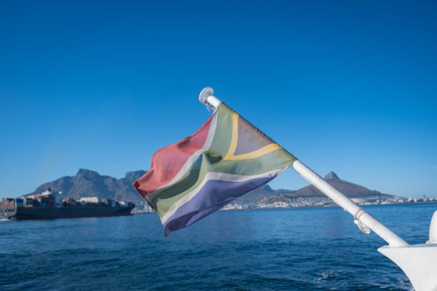 A South African Flag waving off the back of a nautical vessel in the Cape Town Harbor with Table Mountain in the background