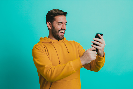 Handsome young man wearing a yellow hoodie having a video call on a smartphone, studio shoot in front of a blue background