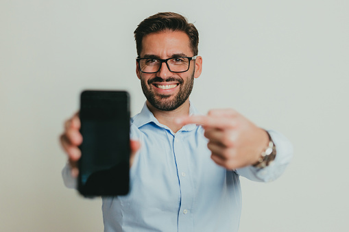 Portrait of a handsome young man wearing a button-down shirt and eyeglasses showing a smartphone screen, studio shoot in front of white background