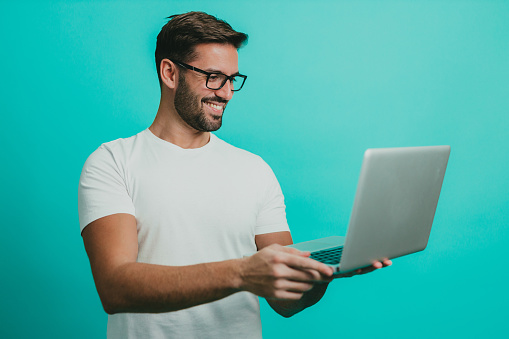 Young man wearing eyeglasses having a video call on a laptop, studio shoot in front of blue background