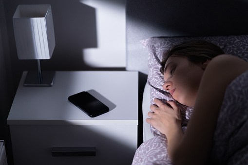 Sleeping woman in bed with phone on table at night. Cellphone on nightstand. Smartphone with silent mode, mute or turned off next to resting person. Dark home bedroom.