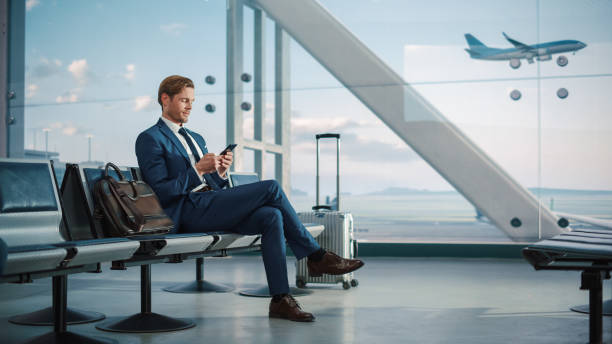 terminal del aeropuerto: hombre de negocios usa teléfono inteligente, espera un vuelo, hace negocios electrónicos, envía datos de comercio electrónico. hombre viajero trabajo remoto en línea en el teléfono móvil en la sala de embarque del centro de  - commercial airplane airport arrival departure board business travel fotografías e imágenes de stock