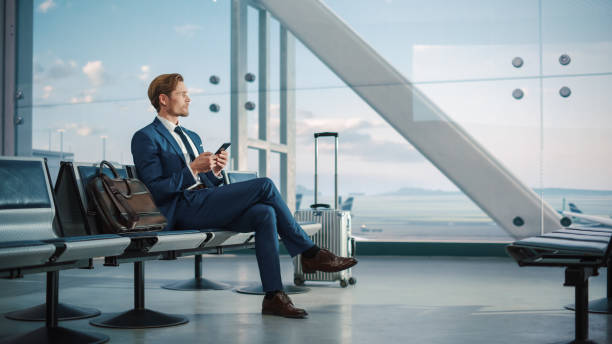 airport terminal: businessman uses smartphone, waiting for a flight, doing e-business, sending e-commerce data. traveling man remote work online on mobile phone in boarding lounge of airline hub - arrival departure board travel business travel people traveling imagens e fotografias de stock