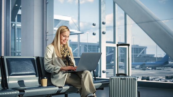 Airport Terminal: Smiling Woman Waits for Flight, Uses Laptop, Browse Internet, does e-Business, Online Shopping. Traveling Female Remote Work Online on Computer in a Boarding Lounge of Airline Hub