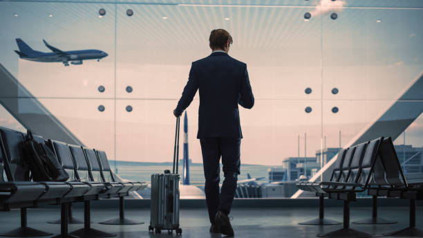 istock Airport Terminal: Businessman with Rolling Suitcase Walks, Uses Smartphone App for e-Business. Back View Silhouette of Traveling Man Waits for Flight in Boarding Lounge of Airline Hub with Airplanes 1387222428