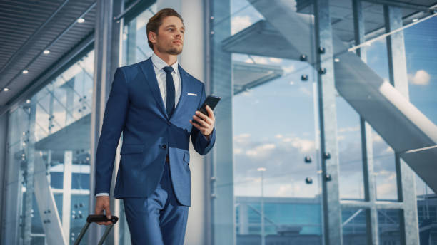 istock Airport Terminal: Businessman Walks to His Flight Gates, Uses Smartphone, Doing e-Business, Browsing the Internet with App. Digital Entrepreneur Remote Work Online While Traveling Through Airline Hub 1387222450