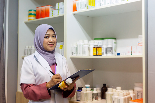 Portrait of female muslim pharmacist wearing lab coat having medicine bottle and writing equipment in medical store.