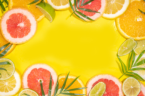 Colorful flatlay of citrus fruit slices and tropical palm leaves. Orange, grapefruit, lemon, lime bright high-colored slices on yellow background. Summer holiday background top view