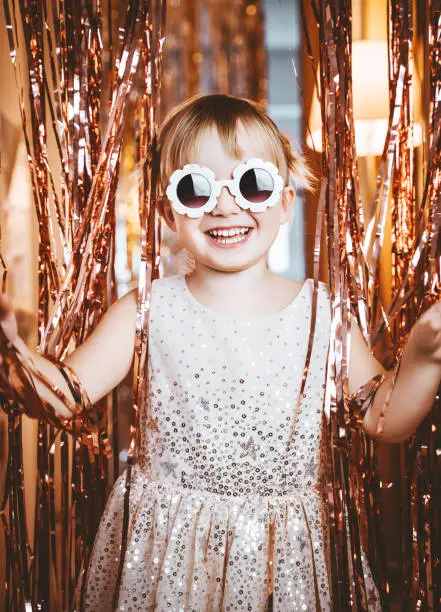 Photo of Portrait of happy little girl in dress and sunglasses having fun on the festive background. Kids birthday or fancy dress party, disco music or New Year. Celebration and Holiday concept.