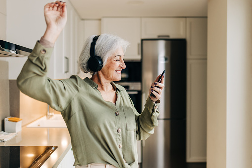 Happy senior woman dancing to her favourite music while wearing wireless headphones. Cheerful elderly woman having a good time while playing music on her smartphone.