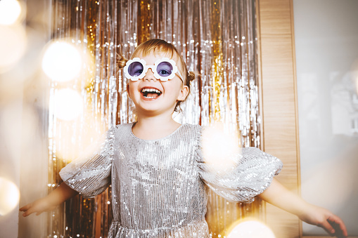 Happy little stylish girl in shiny dress having fun. Festive background with foil curtain decorations for kids birthday or fancy dress party, disco music or New Year. Celebration and Holiday concept.
