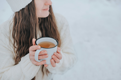 A woman in a white knitted sweater holds a white mug with a hot drink in her hands. Outside in winter.