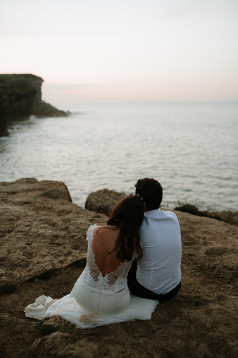 Rear view of a just married couple sitting by the sea at sunset