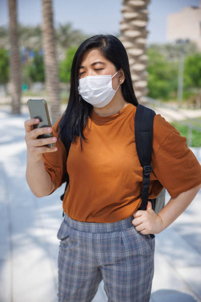 istock Young woman with protective face mask in palm tree alley using mobile phone 1387265241
