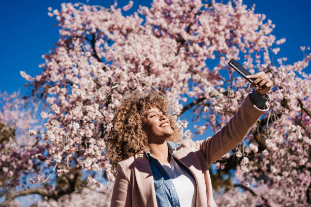 smiling hispanic woman in park taking picture with mobile phone. Spring flowers background stock photo