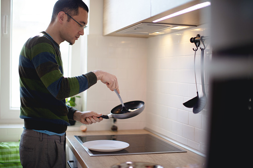 Young man standing by the stove in the kitchen and taking fried eggs from pan with spatula to serve in plate
