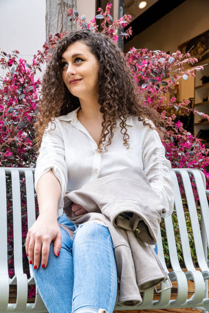 istock Attractive Caucasian female with curly hair posing at camera while sitting on a white bench 1387274732