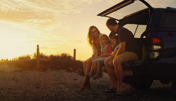 aufnahme einer familie, die am ende ihres fahrzeugs sitzt, während sie am strand sitzt - road trip stock-fotos und bilder