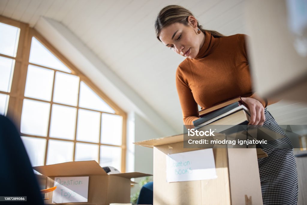 Caring Caucasian young woman packing toys and books for donation into cardboard box Selfless Caucasian female volunteer, packing books and toys for donation into cardboard box Decluttering Stock Photo