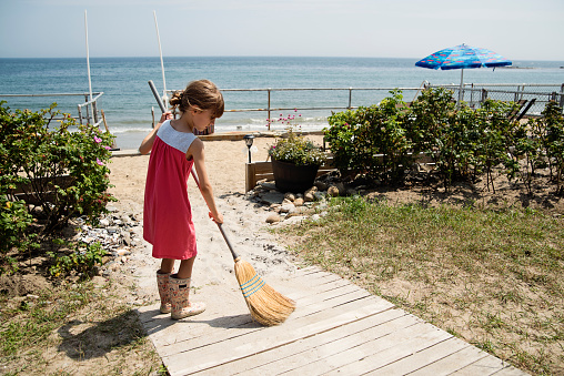 Little girl with broom sweeping sand on a private beach boardwalk. She is 8 year’s old and is wearing a pink dress with rain boots. Horizontal full length outdoors shot wit copy space.