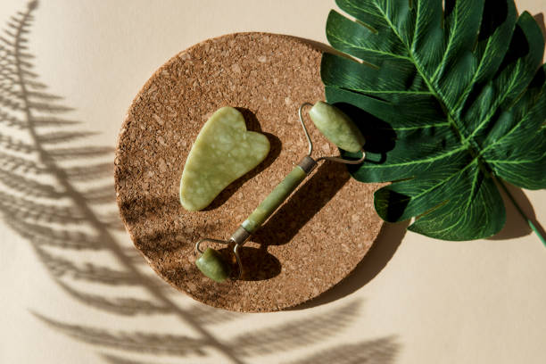 Jade Gua sha scraper and face roller massager on a cork round stand with a monstera leaf stock photo