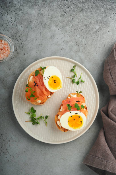 sándwich con deliciosos ingredientes de salmón ahumado, huevos, hierbas y rábano microgreens, semillas de sésamo negro sobre plato blanco sobre fondo de mesa de concreto gris. superalimento saludable de sándwich abierto. vista superior. - smoked salmon cooking copy space food fotografías e imágenes de stock