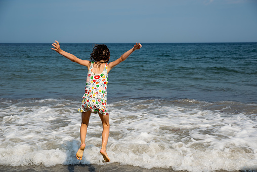Little girl playing in ocean waves. She is 6 year’s old, has curly mid-long hair and is wearing a swimsuit. Horizontal full length outdoors shot with copy space.