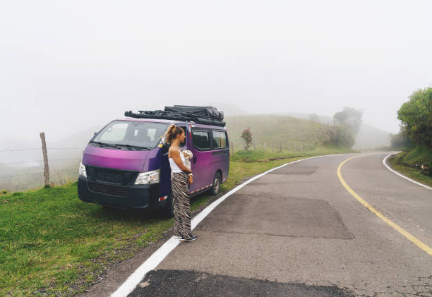 Young Colombian woman standing next to her campervan on the road. Foggy background.