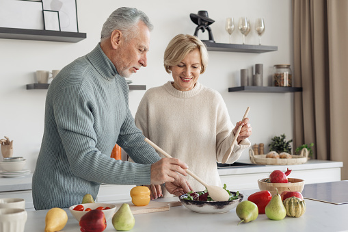 Portrait of smiling senior couple making salad together. Home comfort. Process of cooking lunch together in bright kitchen. Healthy lifestyle