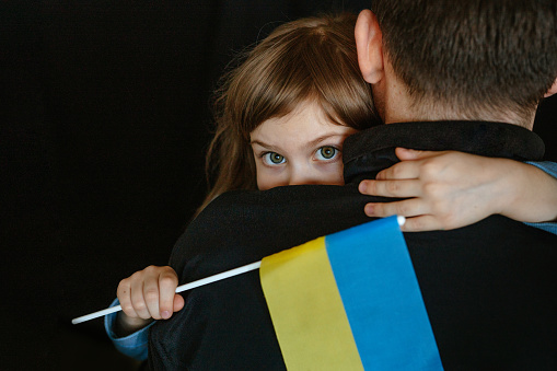 Little girl holding Ukrainian flag hugging her dad standing back. Father with 5 years old daughter on the dark background. Stand with Ukraine. Selective focus on the girl's face.