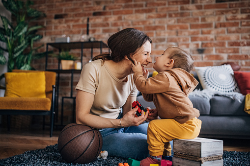 Mother and son playing with toy blocks and different toys  at home in the living room while boy is kissing his mother