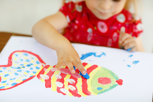 Little creative toddler girl painting with finger colors a fish. Active child having fun with drawing at home, in kindergaten or preschool. Education and distance learning for kids. Creaitve activity
