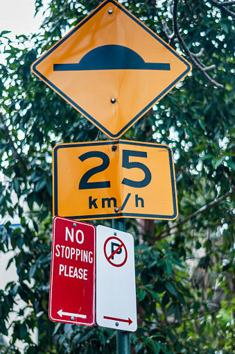 Four road signs on a pole in Australia, No Parking, No Stopping and a speed hump limit sign. The No Stopping sign has had a polite 'PLEASE' sticker added.