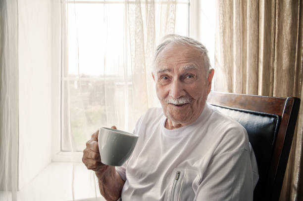 old man is sitting in a chair. A smiling old man is sitting in a chair by the window with a Cup of coffee or tea. portrait, close-up, copy space stock photo